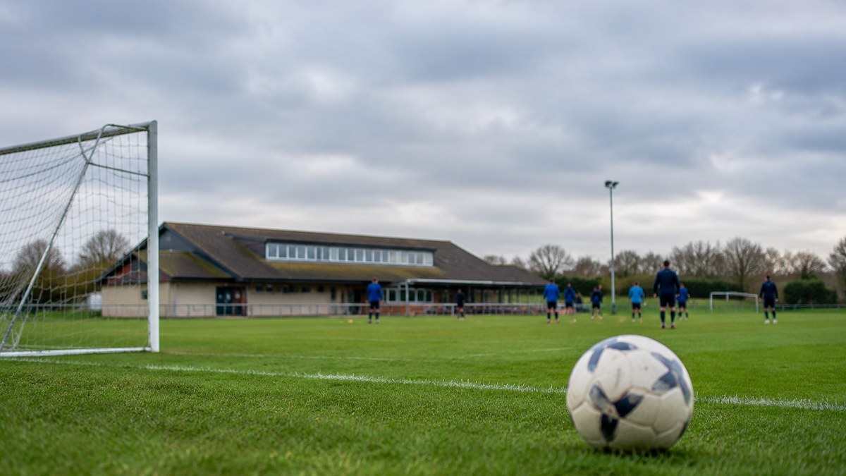 Campo de fútbol amateur en Cambridgeshire preparado para un partido de veteranos, con jugadores anónimos al fondo.